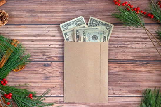 An envelope filled with U.S. dollar bills lies on a wooden surface surrounded by natural holiday decorations, including pine branches, red berries, cinnamon sticks, and a pine cone.