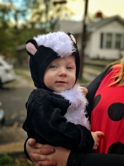 A young child is dressed in an animal costume resembling a skunk, held close by an adult. The costume features black and white fur with small ears. The adult is wearing a red outfit with black polka dots. The background shows an outdoor, residential setting with blurred elements like trees and a house.