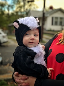 A young child is dressed in an animal costume resembling a skunk, held close by an adult. The costume features black and white fur with small ears. The adult is wearing a red outfit with black polka dots. The background shows an outdoor, residential setting with blurred elements like trees and a house.