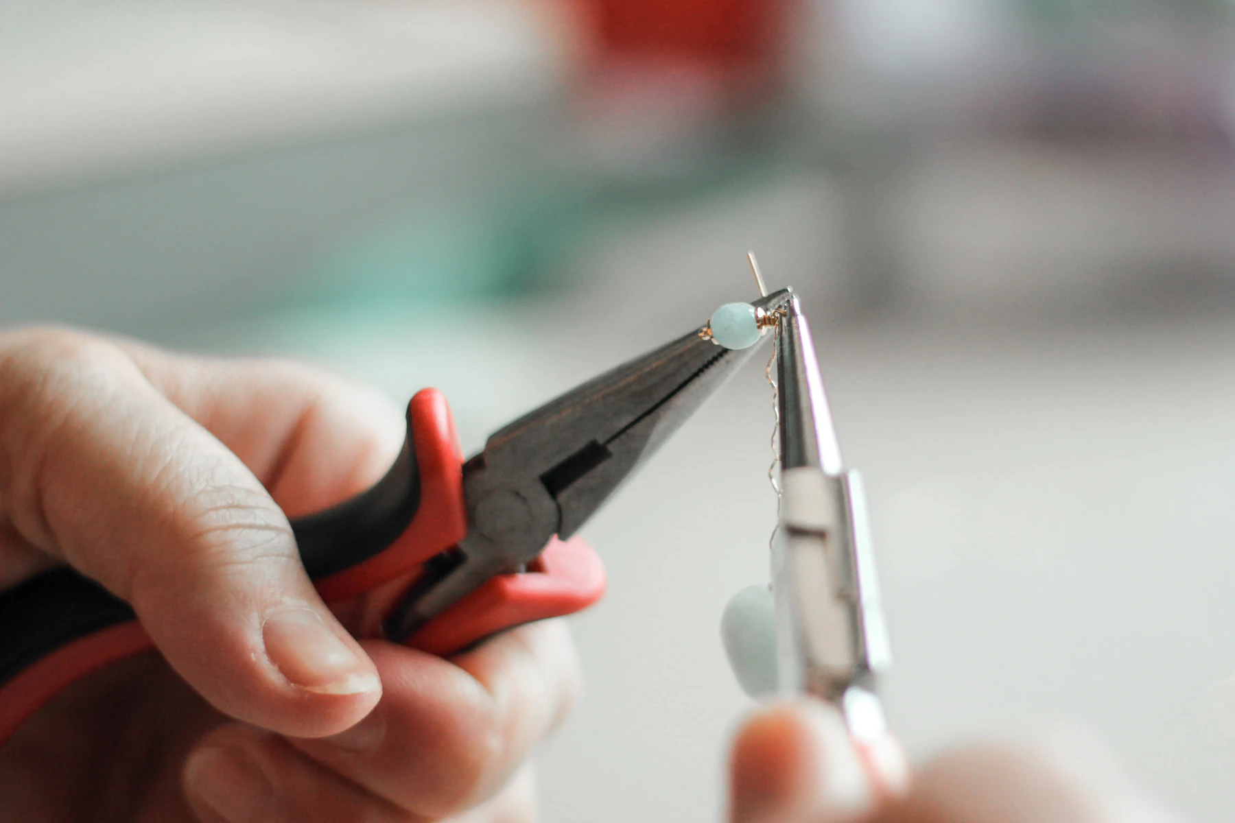 A hand holding red-and-black-handled fine-nose pliers gripping a small metal ring