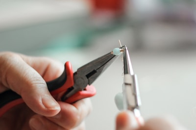 A close-up of hands using two pairs of pliers to work on a small piece of jewelry. One plier holds a small blue bead, while the other adjusts the wire.