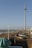 A seaside scene featuring a tall observation tower on the right. The beach is lined with various buildings and colorful beach huts. A promenade runs parallel to the shore, with people strolling along it. The sea is calm with gentle waves, and a clear blue sky stretches above with a few scattered clouds.