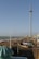 A seaside scene featuring a tall observation tower on the right. The beach is lined with various buildings and colorful beach huts. A promenade runs parallel to the shore, with people strolling along it. The sea is calm with gentle waves, and a clear blue sky stretches above with a few scattered clouds.