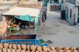 A rustic courtyard area featuring a small cow shelter with a teal canopy attached to a brick wall. Under the shelter, a cow is visible along with farm equipment. In the foreground, a blue tractor trailer with the words 'TRUST IN GOD' is parked, loaded with hay. Near the bottom, several bundles of hay are neatly stacked. The background includes more agricultural tools and structures, along with some residential buildings.