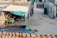 A rustic courtyard area featuring a small cow shelter with a teal canopy attached to a brick wall. Under the shelter, a cow is visible along with farm equipment. In the foreground, a blue tractor trailer with the words 'TRUST IN GOD' is parked, loaded with hay. Near the bottom, several bundles of hay are neatly stacked. The background includes more agricultural tools and structures, along with some residential buildings.