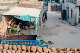 A rustic courtyard area featuring a small cow shelter with a teal canopy attached to a brick wall. Under the shelter, a cow is visible along with farm equipment. In the foreground, a blue tractor trailer with the words 'TRUST IN GOD' is parked, loaded with hay. Near the bottom, several bundles of hay are neatly stacked. The background includes more agricultural tools and structures, along with some residential buildings.