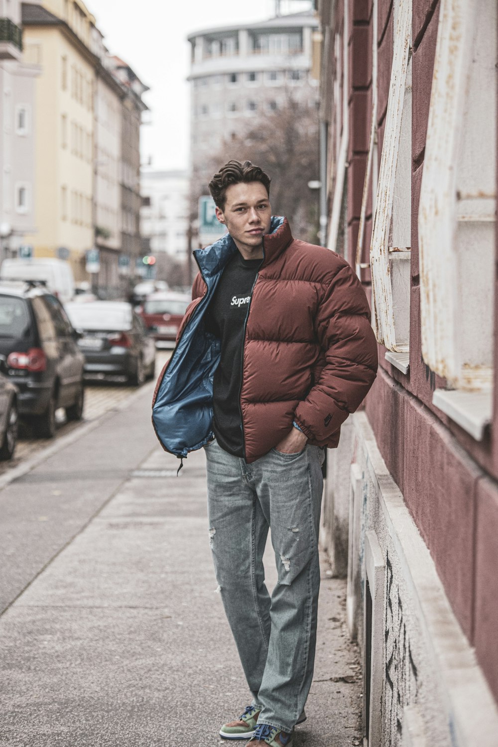 man in red jacket and blue denim jeans standing on sidewalk during daytime