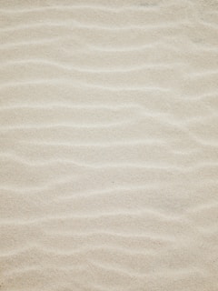 A close-up view of fine sand with subtle wave patterns and ripples creating a textured surface.
