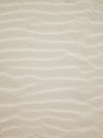 A close-up view of fine sand with subtle wave patterns and ripples creating a textured surface.