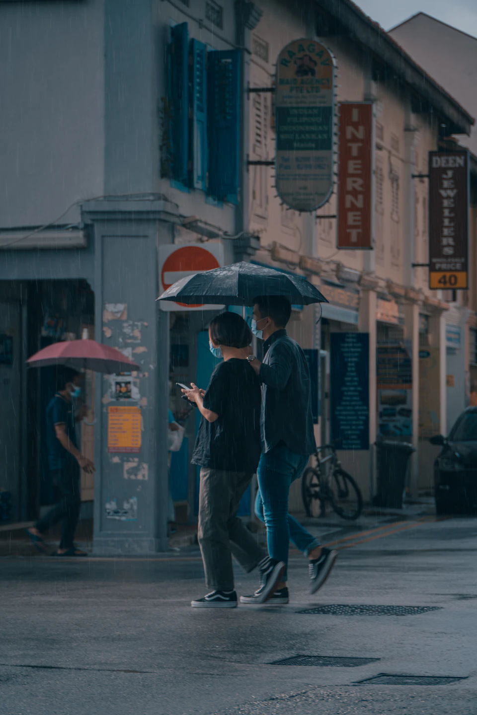 Two people sharing an umbrella crossing a wet street in Little India, Singapore, with shophouse signs behind