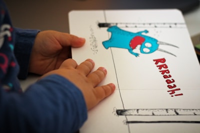 A child's hands are holding a book open to a page with an illustration of a blue cartoon monster with red eyes and an open mouth. The word 'RRRaaah!' is written in red next to the monster. The background is simple, with two vertical lines that resemble tree trunks.