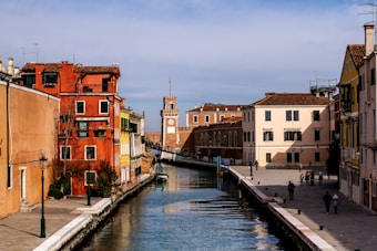 A picturesque canal with clear water runs through a historic Italian town. Colorful buildings in shades of red, yellow, and peach line both sides of the canal. A clock tower is visible in the distance, and a few people are seen walking along the paved pathways that border the water.