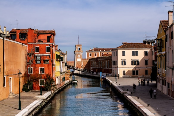 A picturesque canal with clear water runs through a historic Italian town. Colorful buildings in shades of red, yellow, and peach line both sides of the canal. A clock tower is visible in the distance, and a few people are seen walking along the paved pathways that border the water.