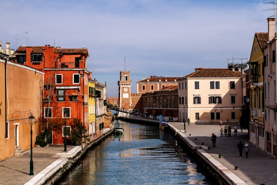 A picturesque canal with clear water runs through a historic Italian town. Colorful buildings in shades of red, yellow, and peach line both sides of the canal. A clock tower is visible in the distance, and a few people are seen walking along the paved pathways that border the water.