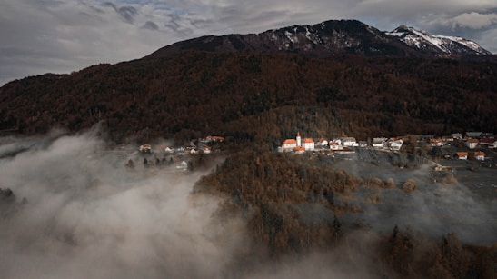 A small village with traditional houses and a church nestled at the base of a forested mountain. Mist and low clouds hover over the landscape, creating an ethereal atmosphere. The mountains in the background are partially covered with snow, adding contrast to the dark green and brown trees.