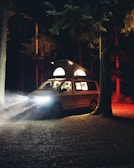 A camper van parked in a forest at night emits light from its elevated roof and headlights, casting an ambient glow on the surrounded trees. The environment is dimly lit with shadows and red hues from the trees providing a dramatic contrast.