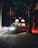 A camper van parked in a forest at night emits light from its elevated roof and headlights, casting an ambient glow on the surrounded trees. The environment is dimly lit with shadows and red hues from the trees providing a dramatic contrast.