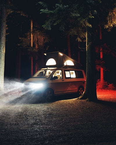 A camper van parked in a forest at night emits light from its elevated roof and headlights, casting an ambient glow on the surrounded trees. The environment is dimly lit with shadows and red hues from the trees providing a dramatic contrast.
