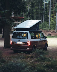 A campervan parked in a lush forest clearing, sunlight filtering through tall trees.