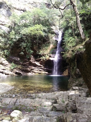 A picturesque waterfall cascades into a serene pool surrounded by lush green foliage and rocky cliffs. Sunlight filters through the trees, creating a tranquil and natural atmosphere.