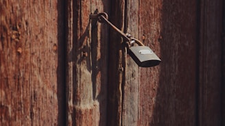 A rustic wooden surface with a weathered appearance, featuring a metallic padlock hanging from a latch. The wood showcases various shades of brown and displays the texture of grain and knots, conveying an aged and worn character.