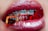 Close-up of lips with shiny red lipstick, revealing braces on the teeth holding translucent red and orange gummy candies.