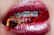 Close-up of lips with shiny red lipstick, revealing braces on the teeth holding translucent red and orange gummy candies.