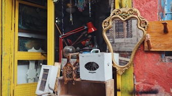 A collection of vintage items is arranged outside a shop with a bright yellow frame door. The scene includes a decorative golden mirror, a red desk lamp, wooden carvings, and various other rustic objects. The surrounding wall is painted in weathered red with patches of blue.