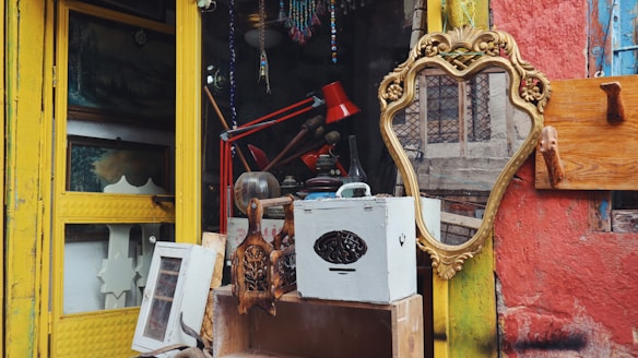 A collection of vintage items is arranged outside a shop with a bright yellow frame door. The scene includes a decorative golden mirror, a red desk lamp, wooden carvings, and various other rustic objects. The surrounding wall is painted in weathered red with patches of blue.
