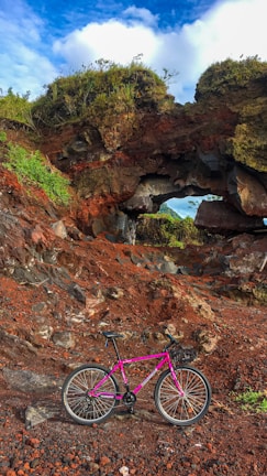 A vibrant pink bicycle is parked on reddish-brown rocky terrain beneath a natural arch formation. The arch is covered with greenery on top, and the sky above shows a mix of clouds and blue. The scene conveys a sense of the outdoors and adventure.