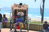 A person sits comfortably in a rickshaw adorned with colorful patterns and beads, with a backdrop of a sunny beach and ocean. There are other people nearby, including a child looking at a phone.