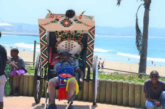 A person sits comfortably in a rickshaw adorned with colorful patterns and beads, with a backdrop of a sunny beach and ocean. There are other people nearby, including a child looking at a phone.