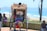 A person sits comfortably in a rickshaw adorned with colorful patterns and beads, with a backdrop of a sunny beach and ocean. There are other people nearby, including a child looking at a phone.