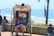 A person sits comfortably in a rickshaw adorned with colorful patterns and beads, with a backdrop of a sunny beach and ocean. There are other people nearby, including a child looking at a phone.