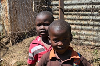 Two young children standing in front of a wire fence with corrugated metal in the background. The child in the foreground is wearing a brown shirt with an orange collar and colorful patches. The child in the background is wearing a red and white striped shirt. Both children are looking towards the camera with neutral expressions.