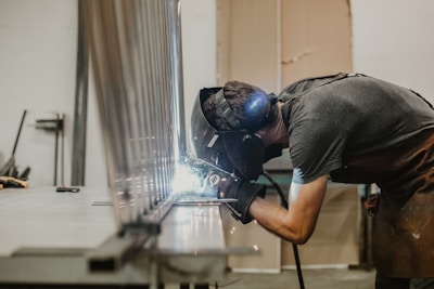 A person wearing a protective welding helmet and gloves is actively welding a metal structure in a workshop. Sparks and bright light are visible as the welding process progresses.