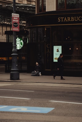 A person sitting on the sidewalk near a Starbucks, with a passerby walking in front. The scene includes a street lamp, a sign in German, and urban surroundings with muted tones.