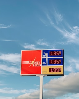 A gas station price sign stands against a backdrop of a blue sky with light clouds. The sign displays prices for regular cash, regular credit, and diesel, with the brand name Energy Fuels prominently featured on a red background.