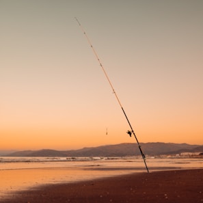 A solitary fishing rod is firmly planted in the wet sand of a tranquil beach. The background features gently rolling waves and distant mountains under a serene orange and blue sky during sunset.
