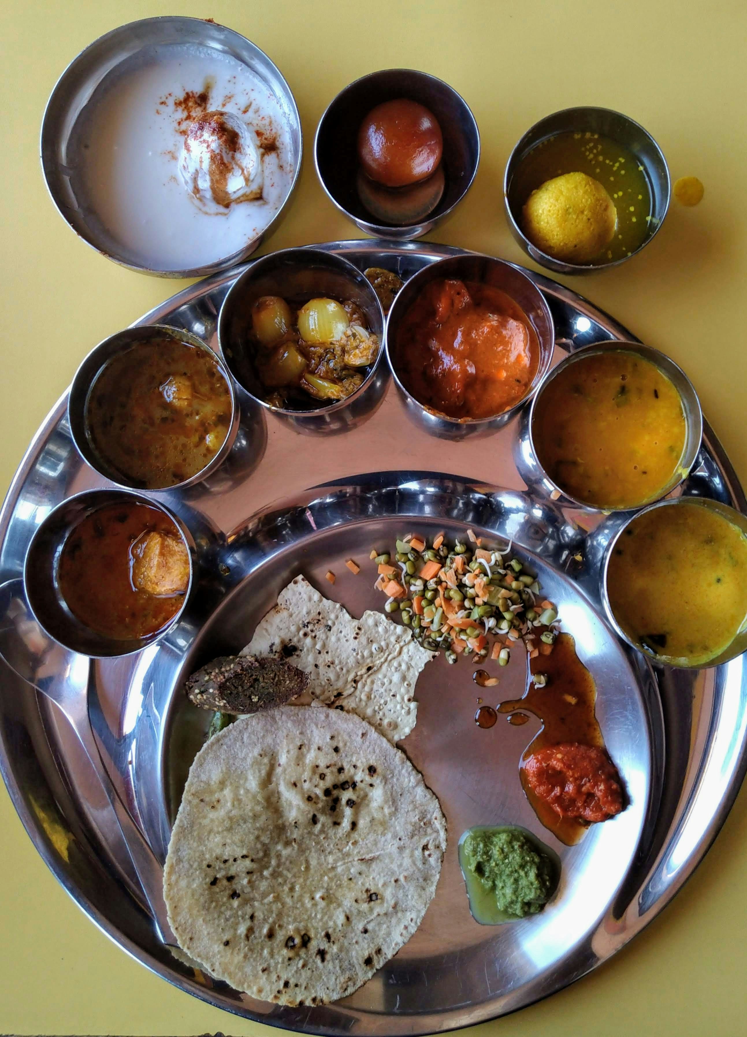 Traditional Rajasthani thali with multiple small bowls of colorful curries, lentils, bread, rice, and sweets arranged in a circular pattern on a silver plate