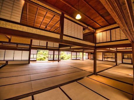 An interior view of a traditional Japanese room featuring tatami mat flooring, wooden beams, and shoji screens. The space is expansive, with a warm, inviting light illuminating the room. The design emphasizes simplicity and harmony, with a subtle blend of natural materials and soft lighting. A small garden is visible through the windows, enhancing the serene atmosphere.