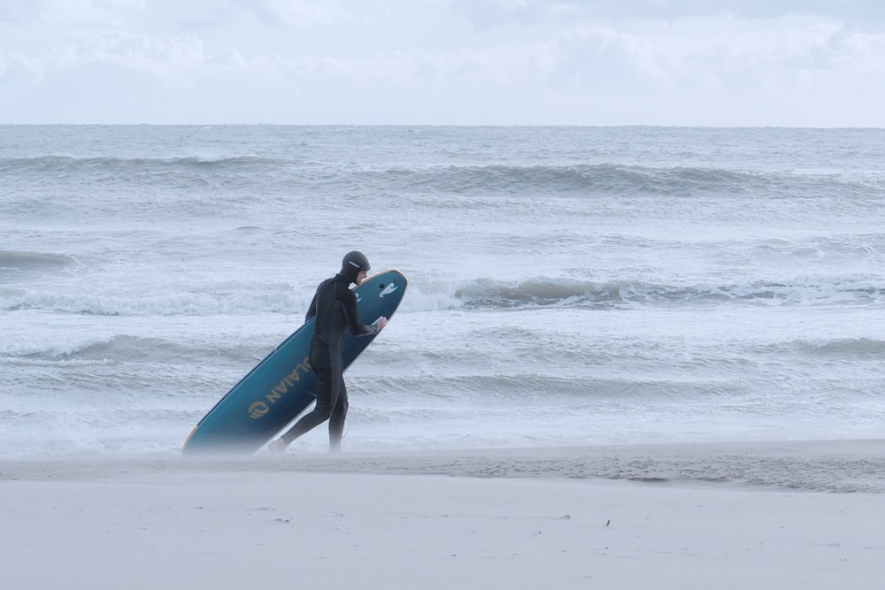 hombre con traje de neopreno azul sosteniendo una tabla de surf azul caminando en la playa durante el día