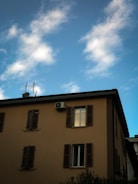 A building with multiple windows and closed shutters is visible against a partially cloudy blue sky. An air conditioning unit is mounted near one of the windows, and antennas are present on the roof.