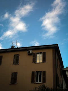 A building with multiple windows and closed shutters is visible against a partially cloudy blue sky. An air conditioning unit is mounted near one of the windows, and antennas are present on the roof.