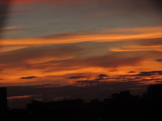 The image displays a sunset or sunrise with vibrant orange, yellow, and dark gray colors streaking across the sky. Dark silhouettes of buildings and possibly trees can be seen at the bottom of the image, adding depth and contrast to the colorful sky.