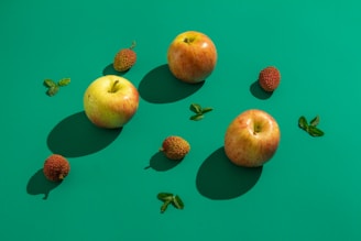 Three apples and several lychee fruits are artistically arranged on a vibrant green background. The apples have a warm, red and yellow hue, while the lychees appear rough-textured and reddish brown. Small sprigs of green mint leaves are scattered between the fruits, adding freshness and contrast.