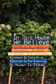 Brightly colored sign with various affirmations is placed in a lush, green garden. The sign reads: 'In This House We Believe,' followed by statements such as 'Black Lives Matter,' 'Science is Real,' 'Love is Love,' 'Kindness is Everything,' 'Feminism is for Everyone,' and 'No Human is Illegal.' The sign is divided into multi-colored sections, each bearing a different phrase.