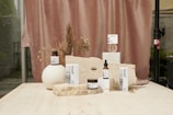 A collection of skincare products displayed on a light beige stone surface. The background features a soft, muted pink curtain with dried floral arrangements, adding an organic and natural element. The products are arranged in an aesthetically pleasing manner, with variations in height and texture for visual interest.