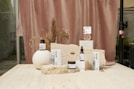 A collection of skincare products displayed on a light beige stone surface. The background features a soft, muted pink curtain with dried floral arrangements, adding an organic and natural element. The products are arranged in an aesthetically pleasing manner, with variations in height and texture for visual interest.
