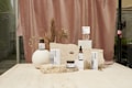 A collection of skincare products displayed on a light beige stone surface. The background features a soft, muted pink curtain with dried floral arrangements, adding an organic and natural element. The products are arranged in an aesthetically pleasing manner, with variations in height and texture for visual interest.